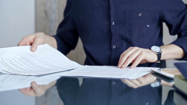 Businessman in dark blue t-shirt analyzing document at desk. Close-up of a professional auditor or lawyer reviewing a lengthy paper report in office setting. Business people concept.